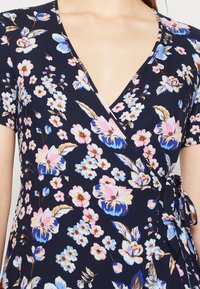 Robe portefeuille marine à manches courtes, ornée de motifs floraux bleus, roses et beiges, nouée à la taille, présentée sur le haut du torse d'une personne.