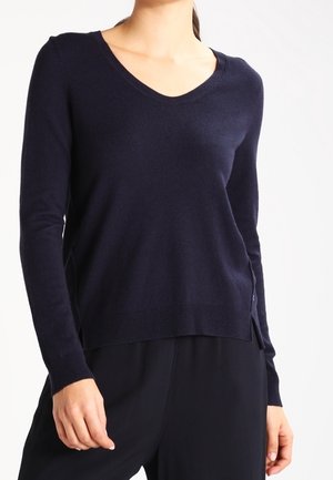 Femme portant un pull bleu marine à col en V et manches longues, ainsi qu'un pantalon noir, debout devant un fond blanc.