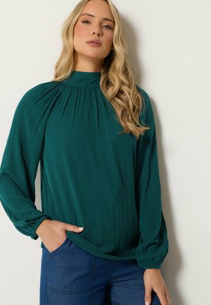 Vrouw met lang blond haar die een donkergroene blouse met hoge hals en geplooide mouwen draagt en blauwe jeans, staand tegen een effen muur.