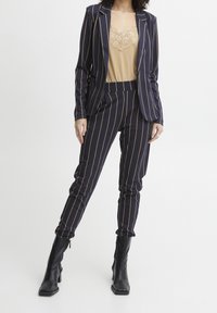 Marinblå randig kostym med figurnära blazer och tapered byxor, kombinerad med en beige topp med spetskant. Svarta ankelskor fulländar looken.