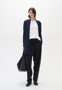 Marine cardigan over en hvit skjorte, kombinert med høyt liv svarte bukser og svarte leiligheter. Modellen holder en stor svart tote-veske.