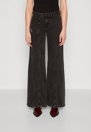 Person trägt hoch taillierte, weit geschnittene schwarze Denim-Jeans mit großen Fronttaschen und burgunderfarbene spitze Stiefel, steht vor einem schlichten Hintergrund.