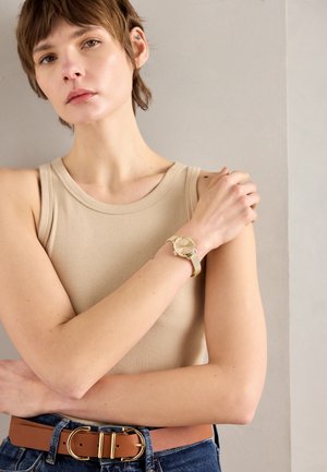 Vrouw in beige tanktop met een gouden horloge en een bruine riem, met gekruiste armen en recht vooruit kijkend tegen een lichtgrijze muurachtergrond.