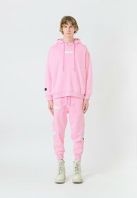 Sudadera rosa y joggers en material suave, con un parche frontal y acentos bordados. Modelo vistiendo botas de combate beige.