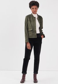 Olivgrön faux suede-jacka med hög krage, framfickor och knappstängning, kombinerad med svarta kordflorsbyxor och bruna ankelstövlar.