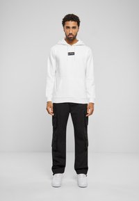 Haut à capuche blanc avec une étiquette noire, poche avant. Assorti avec un pantalon cargo noir avec des poches latérales, porté avec des baskets blanches.
