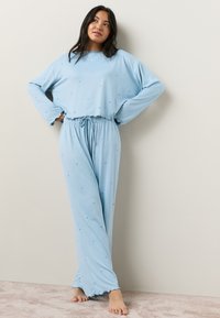 Lyseblåt pyjamasæt med en løstsiddende top med lange ærmer og en snor i taljen. Dekoreret med små stjernebilleder.