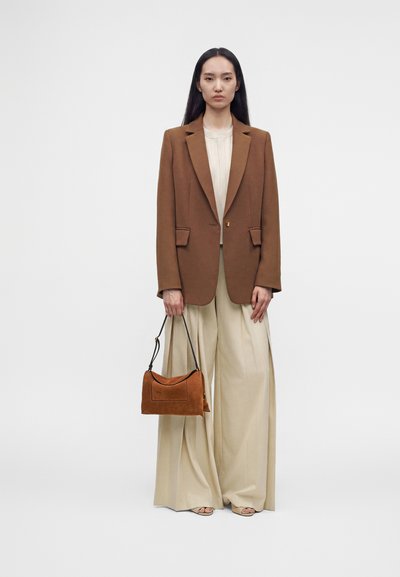 Femme portant un blazer marron, un pantalon beige à jambes larges, tenant un sac à main marron avec une sangle noire, debout devant un fond blanc uni.