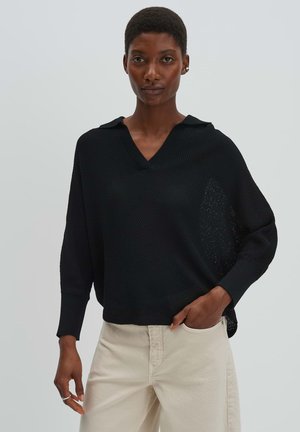 Frau trägt einen locker sitzenden schwarzen strukturierten Pullover mit Kragen und hellbeige Hose, steht mit einer Hand in der Tasche vor einem einfarbigen Hintergrund.