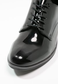 Chaussure habillée en cuir verni noir avec un bout pointu, un laçage et une texture lisse. Présente des coutures discrètes le long des bords.