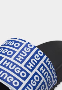 Μαύρο σανδάλι με λουρί σε μπλε και λευκό χρώμα, διακοσμημένο με επαναλαμβανόμενο μοτίβο λογότυπου "HUGO".