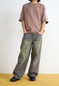 Person som står med händerna i fickorna, iklädd en brun Adidas T-shirt och baggy grå jeans med grönaktig försvagning, svarta skor på en gul golv.