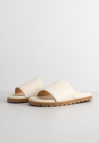 Beige slide sandals made of smooth leather with a wide strap and a flat rubber sole. Features a textured footbed and minimal branding.