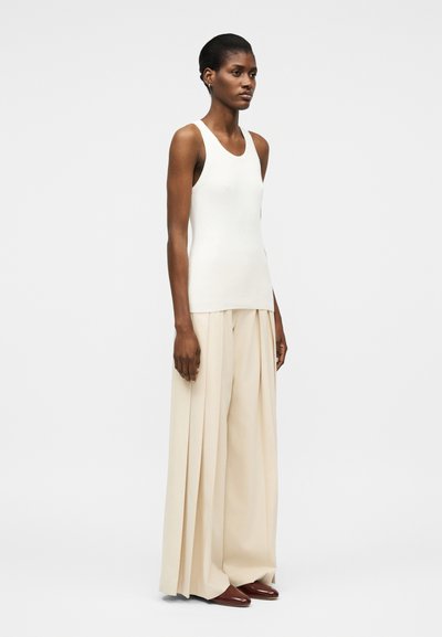 Γυναίκα όρθια με λευκό αμάνικο τοπ, φαρδιά μπεζ πιετέ παντελόνια και καφέ μυτερά παπούτσια σε απλό φόντο.