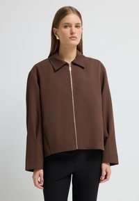 Brun cropped jakke med lynlås, spidse krave og lange ærmer. Stoffet ser tekstureret ud med en glat overflade.