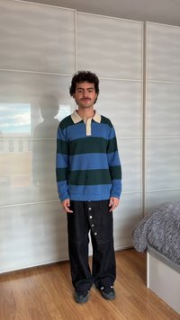 Jonge man met kort krullend haar en snor, draagt een blauw-groen gestreept shirt met lange mouwen, zwarte broek met knopen en zwarte schoenen, staat binnen.