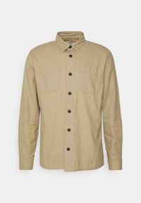 Camicia a maniche lunghe di colore tan realizzata in un tessuto testurizzato. Presenta una chiusura frontale con bottoni, due tasche sul petto e un colletto classico. Bottoni scuri valorizzano il design.