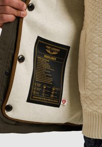 Zelená bunda s krémovým fleecovým podšíváním a zlatou štítkem. Má černé knoflíky, podrobné pokyny k péči a více kapes.