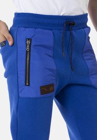 Blaue Jogginghosen aus strukturiertem Stoff, mit einer Reißverschlusstasche und einem Logo-Patch. Der Bund ist elastisch und mit einem Kordelzug zur Anpassung ausgestattet.