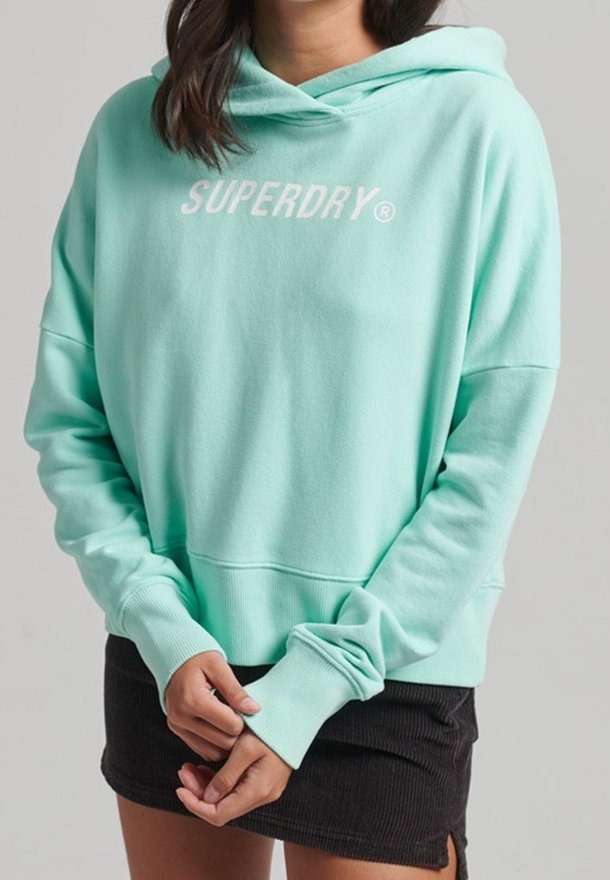 Femme portant un sweat à capuche Superdry turquoise clair à manches longues et une mini-jupe noire sur un fond gris uni.