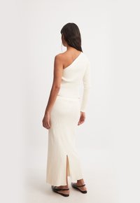 Cremige, gerippte Zweiteiler-Kleid mit einem asymmetrischen Oberteil mit einem langen Ärmel und einem knielangen Rock mit einem seitlichen Schlitz. Flache Sandalen getragen.