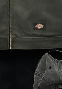 Μαύρο-πράσινο μπουφάν με φερμουάρ από μέταλλο και κοντά στον φθαρμένο γκρι καπέλο μπέιζμπολ βρίσκεται ένα λάβαρο της Dickies σε μαύρη επιφάνεια.