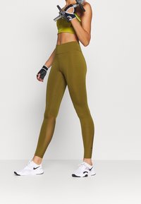 Olive green legginsy z wysokim stanem z panelami siatkowymi, w połączeniu z dopasowanym stanikiem sportowym. Czarno-białe buty sportowe. Hantle w ręku.