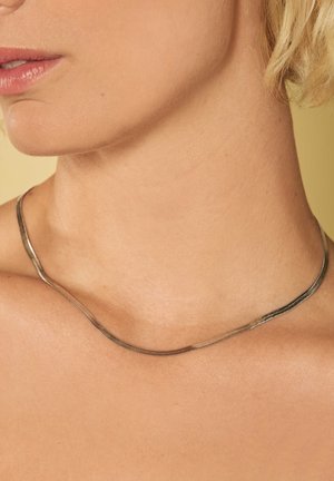 Collier en chaîne serpent en argent, au design lisse et plat, épousant le contour du cou, finition métallique brillante, apparence minimaliste et épurée.
