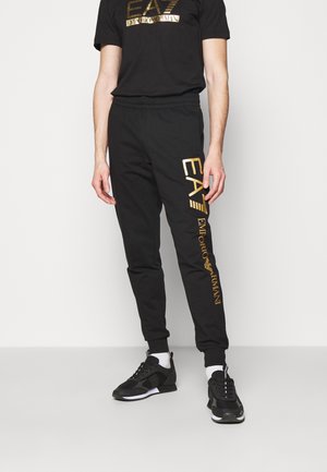 Mężczyzna noszący czarną koszulkę z logo Emporio Armani EA7 oraz pasujące dresy z złotymi literami, do tego czarno-białe sneakersy.