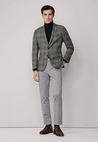 Grå rutig kavaj med snedslåda, dragen över en mörk polotröja. Ljusgrå byxor och bruna läderskor kompletterar outfiten.