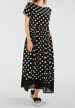 Femme portant une robe longue noire à manches courtes avec des pois blancs et des sandales compensées noires avec des nœuds.