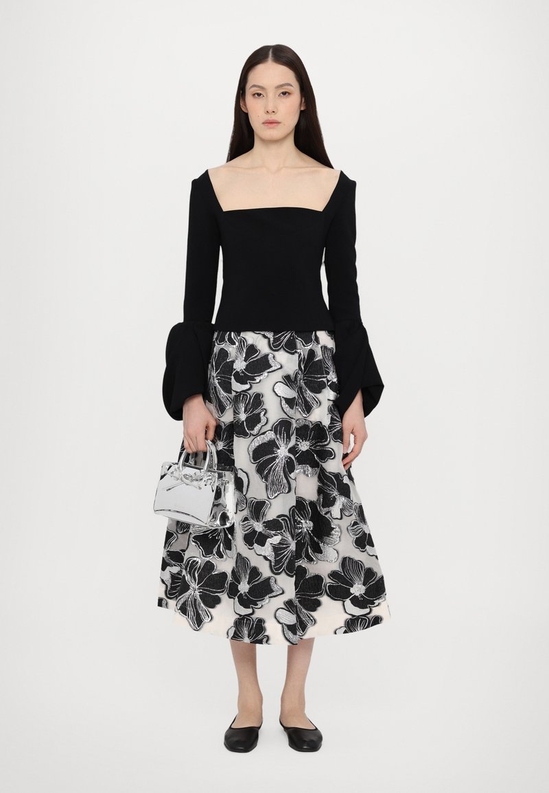 Maglietta nera a maniche lunghe con scollo quadrato, abbinata a una gonna midi stampata floreale in bianco e nero. Gli accessori includono una piccola borsa a tracolla argentata e ballerine nere.