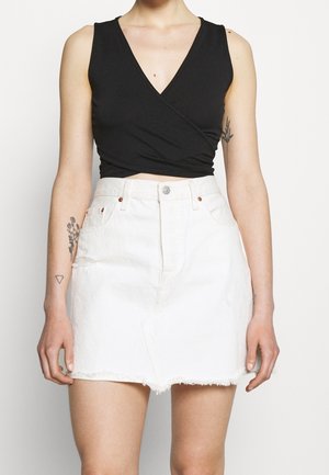 Kobieta w czarnym topie typu wrap bez rękawów oraz w białej, dżinsowej mini spódniczce z wysokim stanem i postrzępionym wykończeniem.