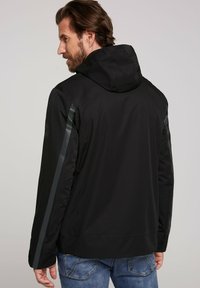 Giacca con cappuccio nera dal finish liscio, con strisce laterali riflettenti. Include un discreto dettaglio del logo sulla spalla.
