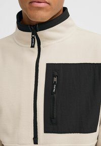 Chaqueta de forro polar en color crema con un bolsillo de nylon negro en el pecho. Dispone de cierre de cremallera y un cuello en contraste. Tejido texturizado con acentos lisos.