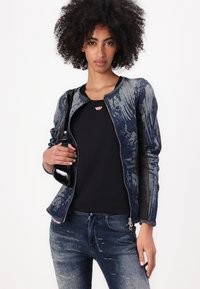 Chaqueta de mezclilla con un diseño azul desgastado y detalles en negro, cuello redondo y cremallera frontal. Combinada con una blusa negra y sosteniendo un bolso negro.