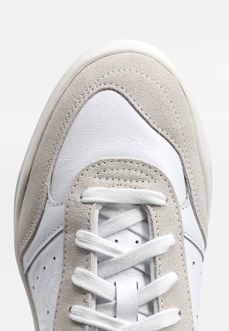 Zapatilla de cuero blanca con acentos de ante gris, puntera redonda, cordones planos y detalles perforados en los lados. Superficies suaves y texturizadas.
