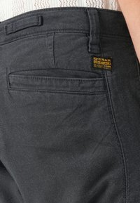 Zadnji žep temno sivo obarvanih džins hlač G-Star RAW, oblečenih z lahkim teksturiranim srajcami, prikazuje blagovno oznako blizu pasu.