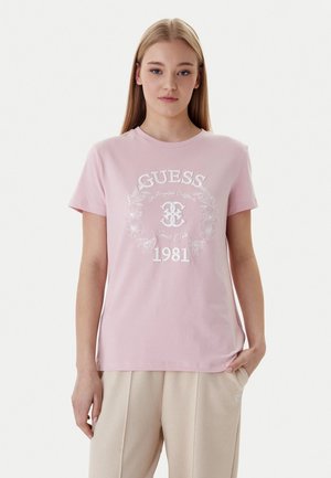 Giovane donna con lunghi capelli biondi che indossa una maglietta rosa chiaro con il logo "GUESS 1981" e pantaloni beige, in piedi con una mano in tasca.