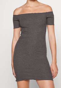 Femme portant une robe ajustée, à côtes, gris foncé, décolletée, avec des manches courtes, se tenant contre un fond clair uni.