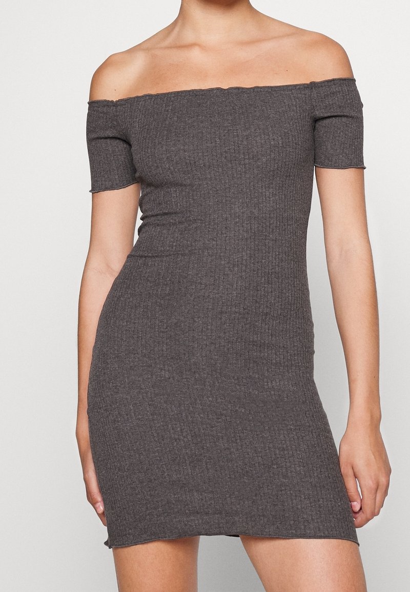 Frau in einem figurbetonten, gerippten, dunkelgrauen, schulterfreien Kleid mit kurzen Ärmeln, die vor einem einfarbigen hellen Hintergrund steht.