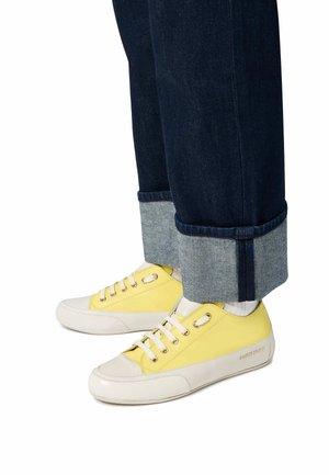 Geel en witte sneakers met een gladde leren bovenkant, ronde neus en metalen veterogen, gecombineerd met donkere denim jeans met opgerolde boorden.