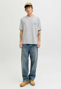 Siv majica iz bombaža s plavim potiskom, ohlapne modre kavbojke in bež delovne čevlje. Dodatki vključujejo zeleni klobuk in srebrno verigo.