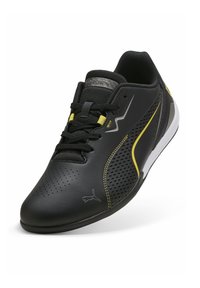 Musta urheilukenkä, jossa on rei'itetty päällinen, keltaiset korostusviivat ja teksturoidut mustat nauhat. Kenkässä on valkoinen pohja ja brändilogolla varustettu merkki.