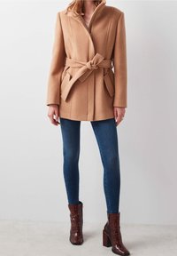 Kameelkleurige wollen jas met een riem in de taille, ritsluiting en zijzakken, gecombineerd met donkerblauwe jeans en bruin gemarmerde enkellaarsjes.