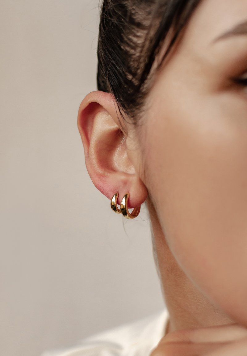 Złote kolczyki w kształcie obręczy z trzema splatającymi się konturami. Gładkie, lustrzane wykończenie; przymocowane do przekłutej małżowiny usznej, podkreślające wzór.
