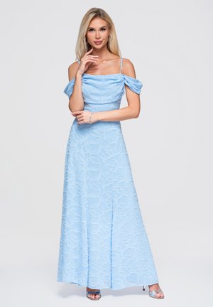 Lyshåret kvinde iført en lyseblå off-shoulder maxikjole med blomsterstruktur og sølvfarvede højhælede sandaler, poserer med en hånd nær ansigtet.