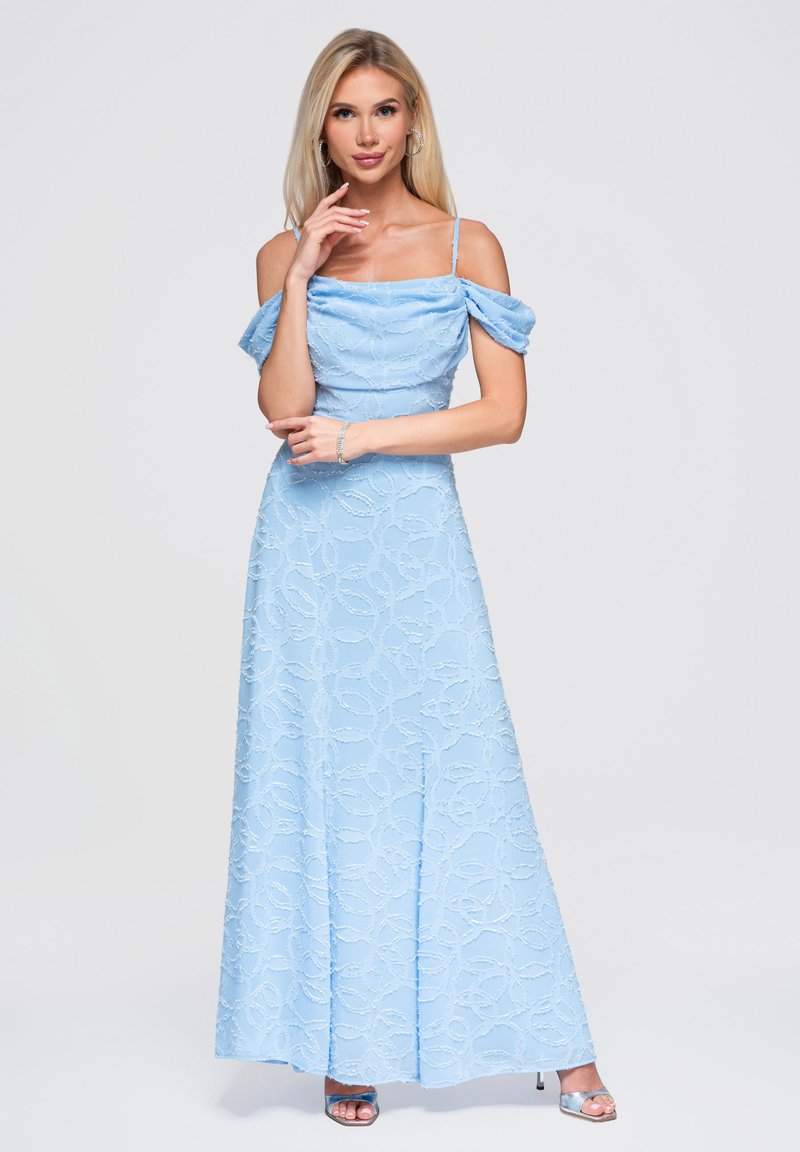 Femme blonde portant une robe maxi bleu clair épaules dénudées avec une texture florale et des sandales argentées à talons hauts, posant avec une main près du visage.