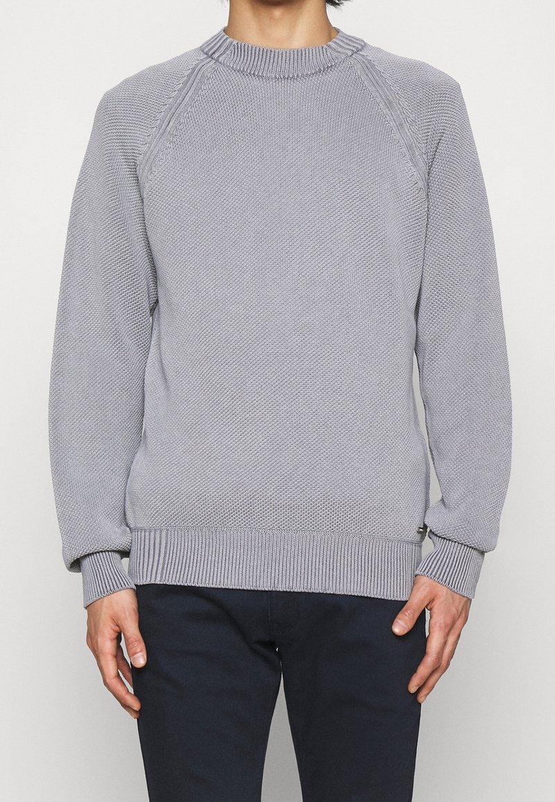Homme portant un pull en maille texturée gris clair avec poignets, ourlet et encolure côtelés, associé à un pantalon foncé sur un fond uni.