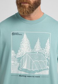 Helles teal T-Shirt mit einem weißen Grafiken eines Zeltes, das zwischen Bäumen steht, und dem Text "Raum zum Umherstreifen schaffen." Weicher Stoff.